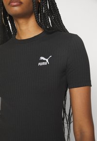 Femme aux cheveux tressés porte un t-shirt PUMA noir côtelé à manches courtes, des créoles dorées et un fond neutre.