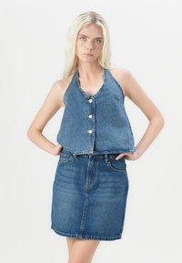 Denim halter top s petimi gumbi, v kombinaciji z denim krilom. Obe kosi sta srednje modri, z gladko teksturo in rahlim bledenjem.