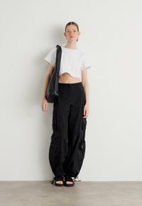 Κοντό λευκό τοπ, μαύρο cargo παντελόνι με τσέπες και μαύρες σαγιονάρες. Το μοντέλο ποζάρει με μία μαύρη τσάντα μπροστά σε έναν άδειο τοίχο.