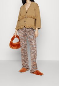 Cardigan brun en tricot avec des détails en franges, associé à un pantalon texturé multicolore et un sac à main orange duveteux. Des chaussons orange sans lacets complètent le look.