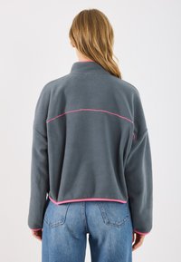 Gray fleece jacket with a high collar and pink piping along the hem and seams, featuring a relaxed fit and dropped shoulders.