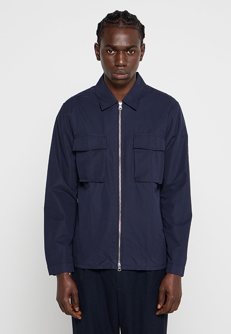 Chaqueta azul marino hecha de tejido ligero, con cremallera frontal, dos bolsillos en el pecho y cuello de punto. Diseño minimalista y textura suave.