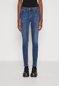 Jeans ajustados de mezclilla azul oscuro, de cintura media, con diseño de cinco bolsillos, costuras en contraste y combinados con zapatos negros.