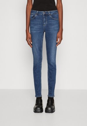 Jeans skinny in denim blu scuro, vita media, con design a cinque tasche, cuciture a contrasto e indossati con scarpe nere.