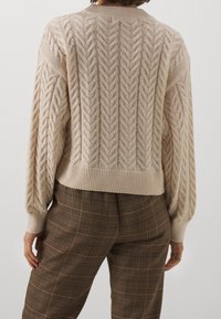 Kabelstickad tröja i ljusbeige med ett texturerat mönster, långa ärmar och ribbad nederkant, i kombination med bruna rutiga byxor.