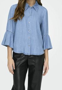 Chemise en denim bleu clair avec un devant à boutons, un col pointu et des manches à volants. Associée à un pantalon en cuir noir, montrant des poches.