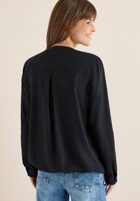 Sort langærmet bluse med en glat tekstur, detalje med plissering bagpå og knapmanchetter, parret med lyseblå jeans.