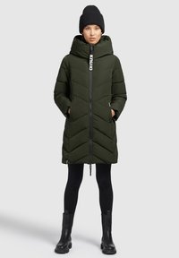 Vadderad olivgrön parka med chevrondesign, dragkedja, avtagbar huva och två sidofickor. Matchad med svarta leggings och stövlar.