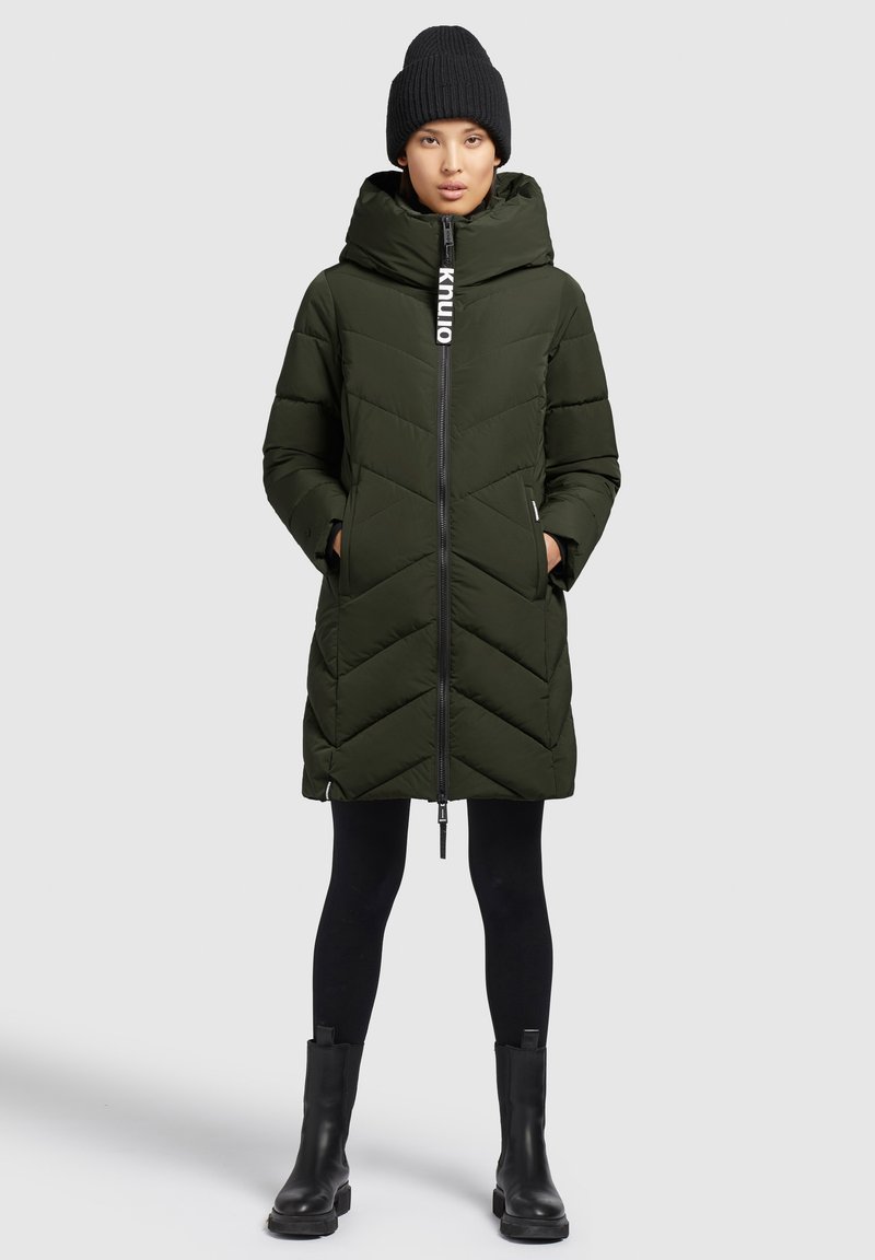 Gepolsterte olivgrüne Parka mit Zickzackmuster, Reißverschluss, abnehmbarer Kapuze und zwei Seitentaschen. Kombiniert mit schwarzen Leggings und Stiefeln.