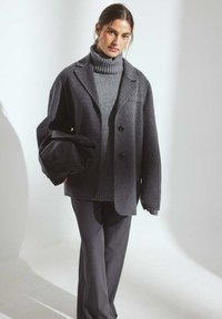 Gray oversized wool blazer with a notched collar, paired with a matching turtleneck sweater and tailored black trousers. Model holds a black bag.