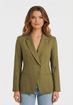 Donna che indossa un blazer verde oliva con un solo bottone sopra jeans blu, in piedi davanti a uno sfondo bianco semplice, rivolta verso la fotocamera.