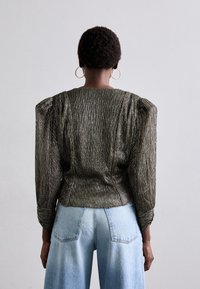 Metaalachtige donkergroene textuurtop met bolle mouwen, getailleerde taille en omgeslagen manchetten, gecombineerd met lichtblauwe wijde jeans.
