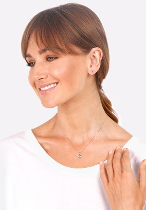 Collier en argent avec un seul pendentif étincelant, porté avec des boucles d'oreilles en argent. Le mannequin a les cheveux raides, châtain clair et porte une chemise blanche.