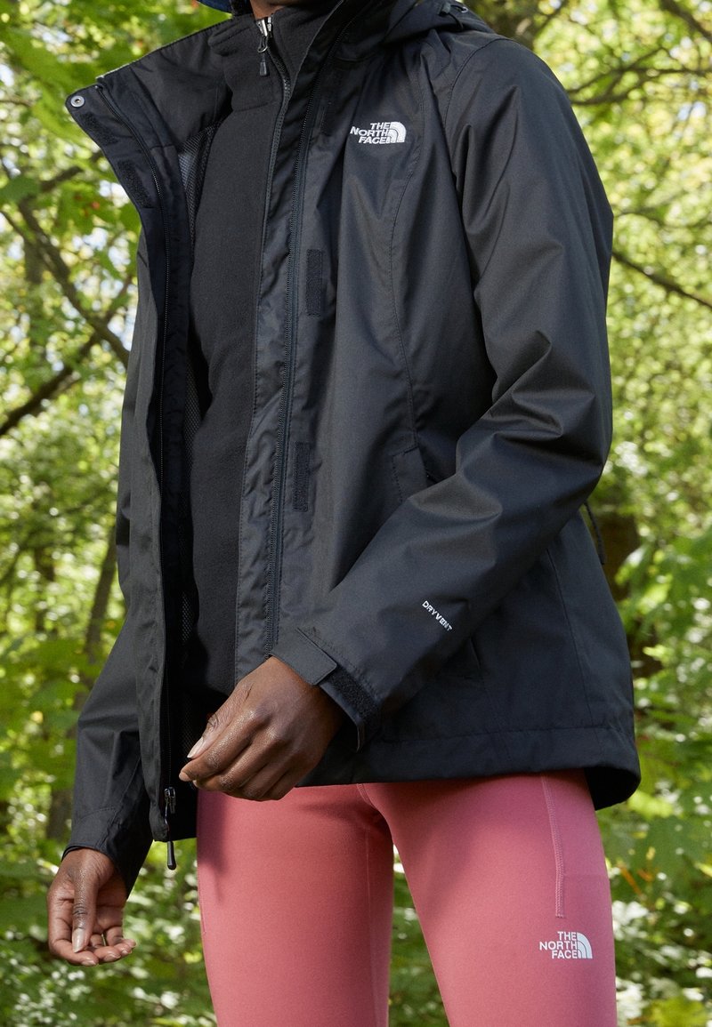Veste noire imperméable avec fermeture éclair à l'avant, capuche ajustable et poignets élastiques. Portée par-dessus un haut noir et associée à des leggings roses.