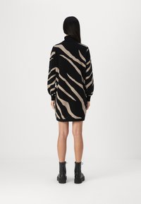 Schwarzes Strickkleid mit beigefarbenen Zebra-Streifen, gerippten Bündchen und einem hohen Halsauschnitt. Das Modell trägt schwarze Knöchelstiefel.