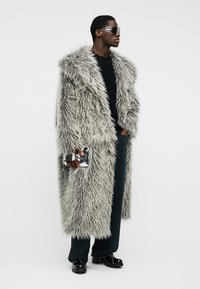 Άνδρας που φοράει μακρύ, ατημέλητο γκρι γούνινο παλτό, μαύρο παντελόνι, μαύρο πουκάμισο, μαύρα παπούτσια, μεγάλα γυαλιά ηλίου και κρατάει μια ασημένια, ανακλαστική τσάντα χειρός.