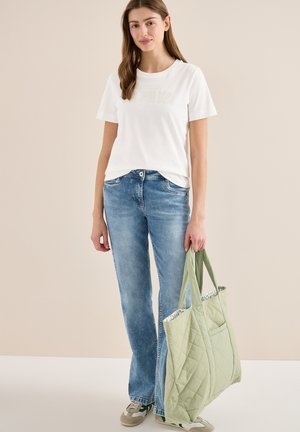 Junge Frau trägt ein weißes T-Shirt und blaue Jeans, hält eine große hellgrüne gesteppten Tragetasche und steht vor einem beigen Hintergrund.