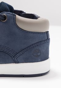 Námořnické modré kožené boty s šedým polstrovaným límcem, bílou gumovou podrážkou a embosovaným logem. Obsahují prošívané akcenty pro příjemnější textura.