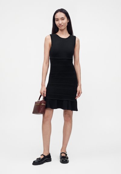 Femme en robe noire sans manches et chaussures noires tenant un petit sac à main marron, debout devant un fond blanc uni.