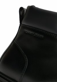 Acercamiento de una bota negra de cuero hasta el tobillo con cuello acolchado y logo en relieve de Tommy Jeans en el panel lateral.