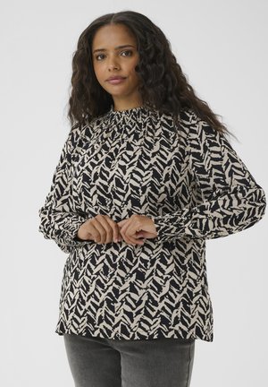 Mujer con cabello oscuro y ondulado que lleva una blusa de manga larga con patrón negro y beige, de pie con las manos entrelazadas al frente, fondo neutro.