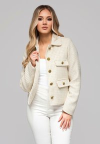 Beżowa marynarka z tweedu o fakturowanym wzorze, z kołnierzem, złotymi guzikami i dwoma przednimi kieszeniami, zestawiona z białą odzieżą.
