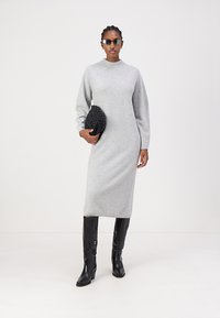 Robe grise tricotée à manches longues avec un col montant, associée à des bottes noires brillantes jusqu'aux genoux et à une pochette texturée noire, sur un fond neutre.