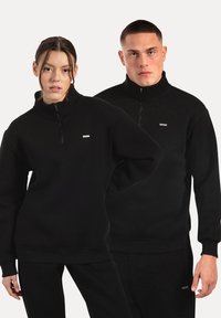 Joven mujer y hombre vestidos con sudaderas negras de media cremallera a juego, de pie uno al lado del otro frente a un fondo neutro.