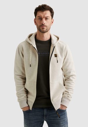 Mann trägt hellbeige Hoodie mit Reißverschluss über dunklem grafischem T-Shirt und blauer Jeans, steht mit Händen in den Taschen vor einfarbigem Hintergrund.
