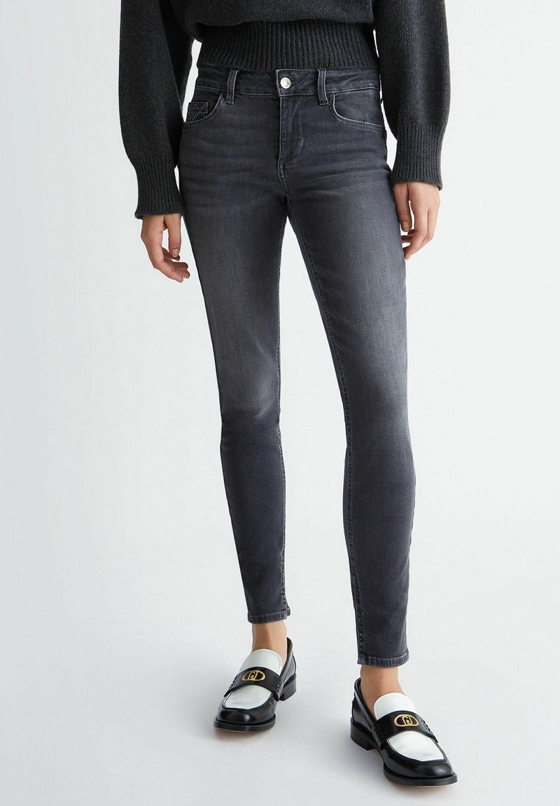 Jeans ajustados en denim gris oscuro con una textura suave y un ligero desgaste. Combinados con mocasines en blanco y negro que presentan acentos dorados.