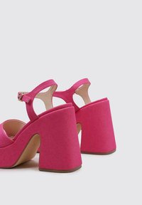 Sandalias de tacón alto de tela rosa con un tacón grueso, correa ajustable en el tobillo y diseño destalonado. Material texturizado y forma curva.