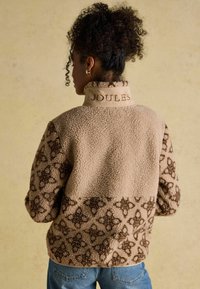 Woman with curly hair wearing a beige patterned fleece jacket with "JOULES" embroidered on the collar, paired with blue jeans.