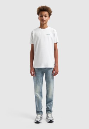 Tienerjongen staat recht voor zich uit kijkend, draagt een wit T-shirt met korte mouwen, lichtblauwe spijkerbroek en witte sneakers tegen een effen achtergrond.