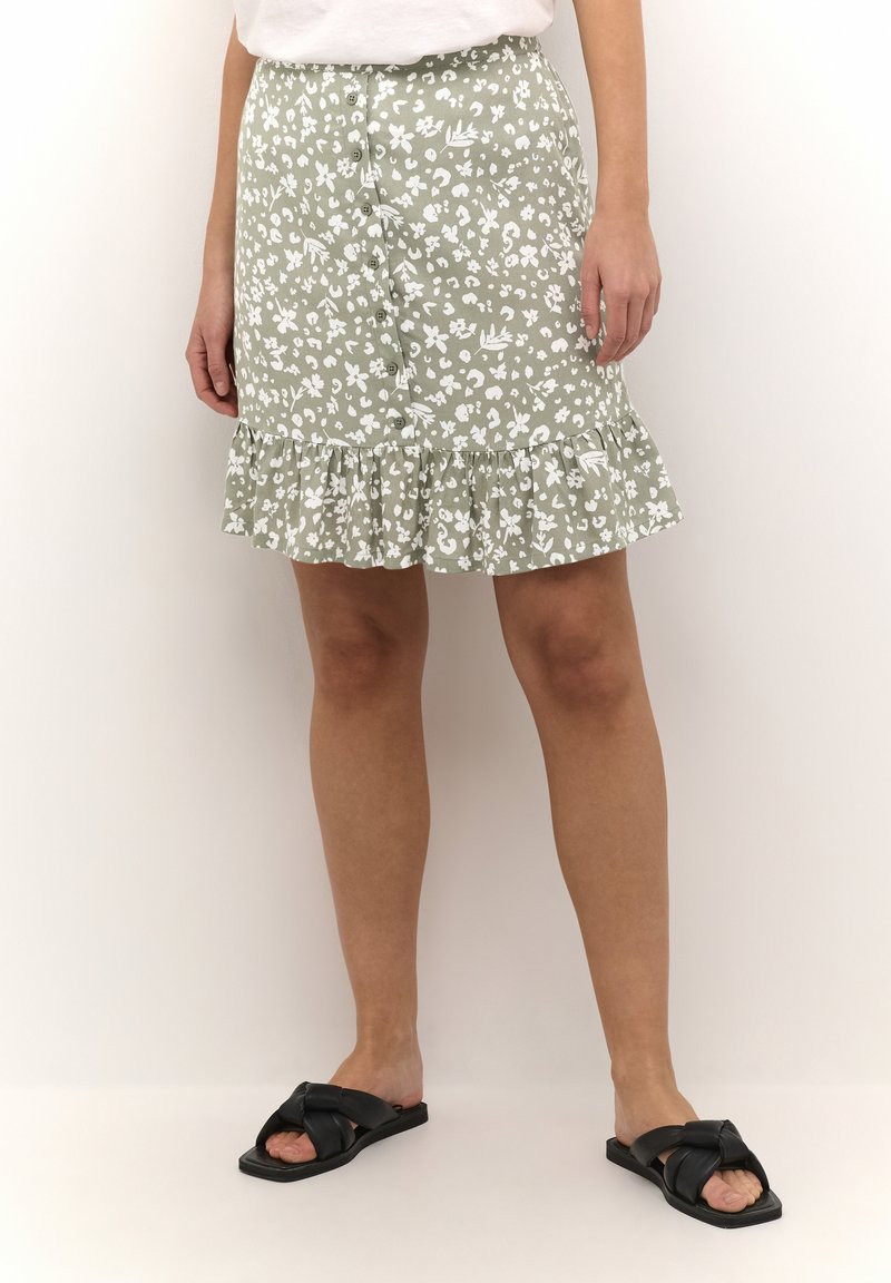 Femme portant une jupe verte fleurie à boutons sur le devant et un ourlet à volants, ainsi que des sandales noires faciles à enfiler, debout devant un fond clair uni.