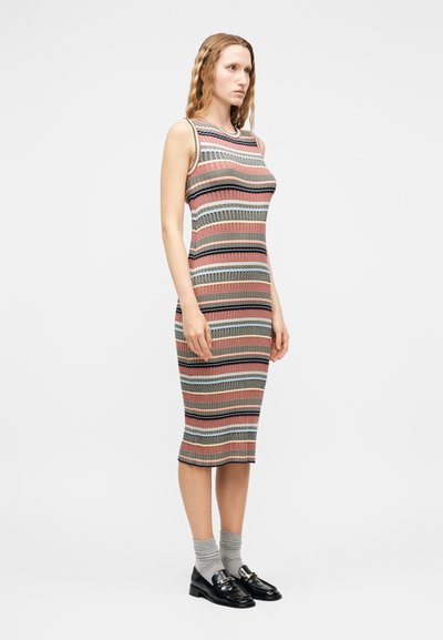 Femme debout portant une robe en maille multicolore à rayures sans manches, longueur genoux, avec des chaussettes grises et des mocassins noirs sur un fond uni.