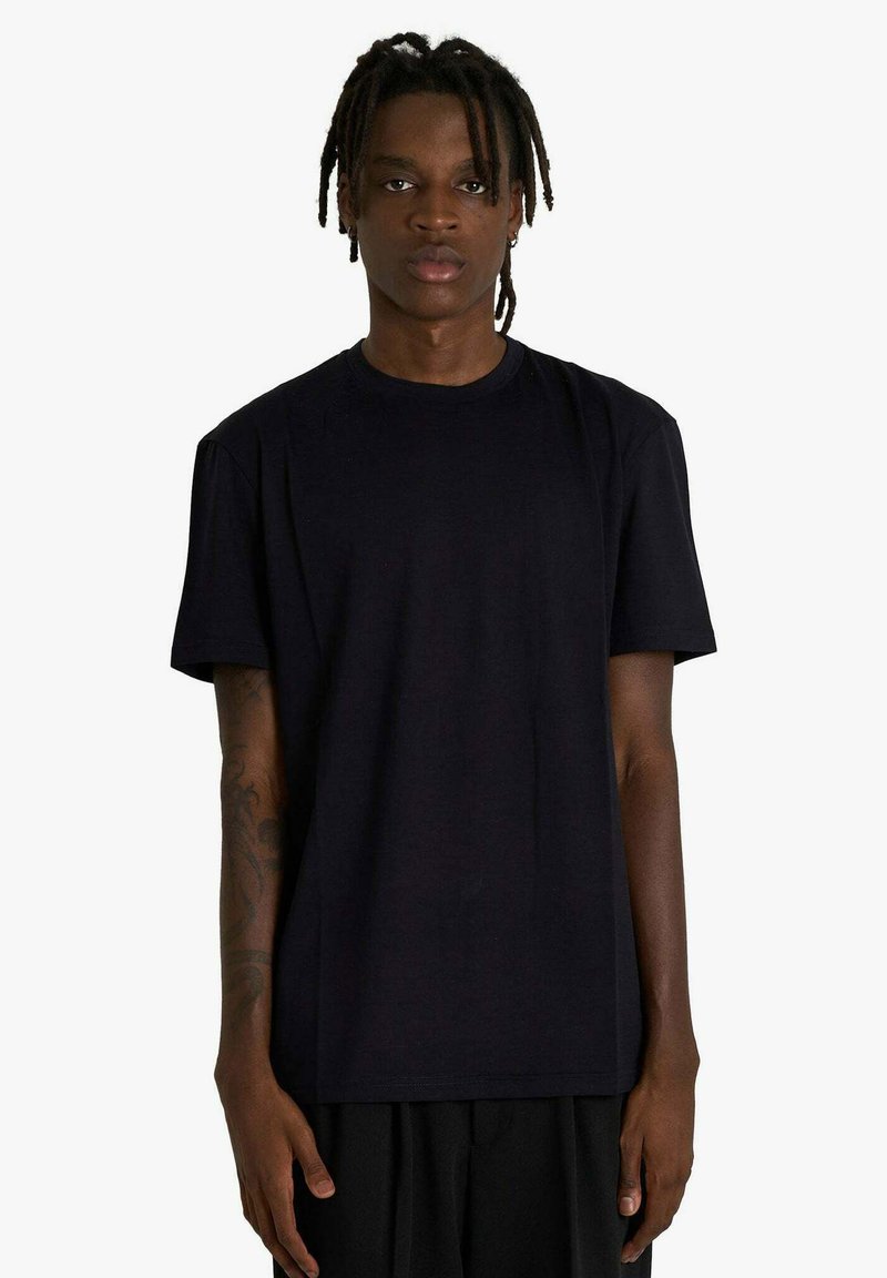 Giovane con dreadlocks che indossa una maglietta nera a maniche corte, in piedi davanti a uno sfondo bianco, con il volto rivolto verso l'obiettivo.