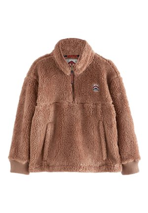 Persona con cabello rizado que lleva una chaqueta con cremallera marrón claro, peluda, Superdry Mountain Sports, sobre una camisa azul con botones.