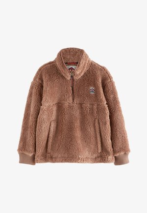 Personne aux cheveux bouclés portant une veste zippée beige et duveteuse Superdry Mountain Sports par-dessus une chemise bleue boutonnée.
