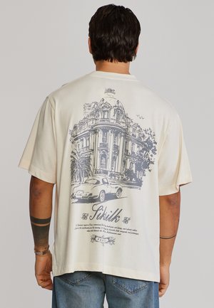 Homme portant un T-shirt crème avec une illustration d'une voiture vintage et d'un bâtiment orné dans le dos, associé à un jean denim bleu.