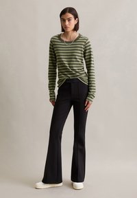 Olijfgroen en zwart gestreepte lange mouwen shirt met een relaxte pasvorm, gecombineerd met zwarte flared broek en witte sneakers, tegen een neutrale achtergrond.