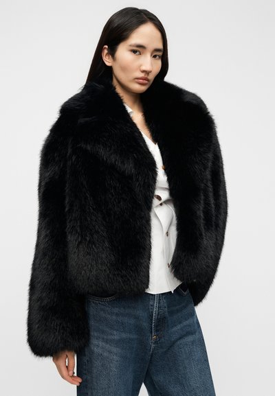 Μαύρο σακάκι από συνθετική γούνα με ψηλό γιακά, κοντό σχέδιο και μακριά μανίκια, φορεμένο πάνω από λευκό πουκάμισο και μπλε τζιν. Μαλακή υφή, χωρίς ορατά σχέδια.