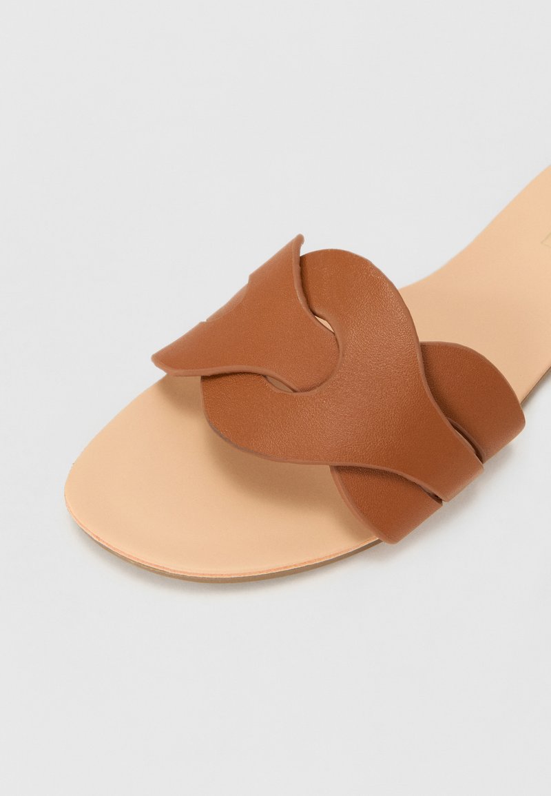 Sandale à glisser en cuir marron avec un design de brides courbées et entrelacées. Semelle intérieure beige avec une texture lisse et une forme minimaliste.