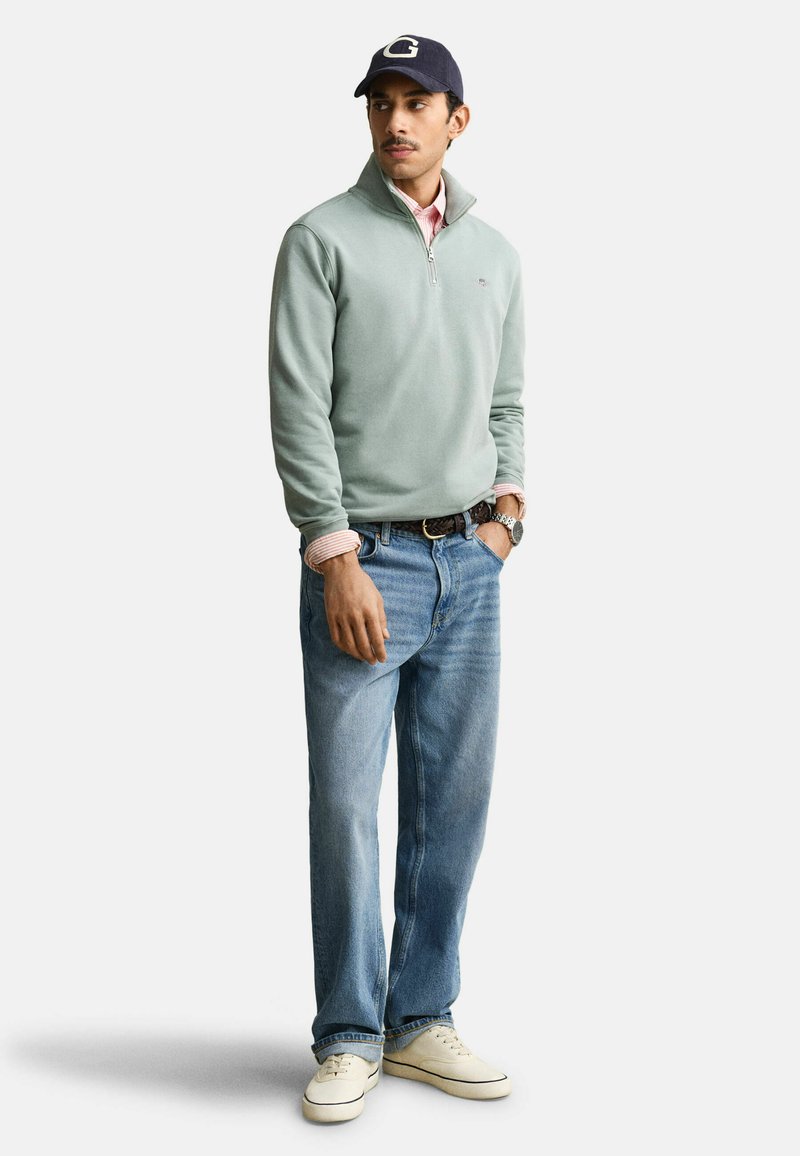 Homme portant un pull léger vert clair à fermeture éclair sur le devant, une chemise rayée, un jean bleu, des baskets blanches, une montre et une casquette bleu marine avec un logo "G", debout, une main dans la poche.