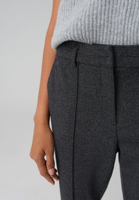 Pantaloni a spina di pesce grigi con una vestibilità sartoriale, dotati di tasche laterali e un maglione testurizzato, mostrati con una mano appoggiata sulla gamba dei pantaloni.