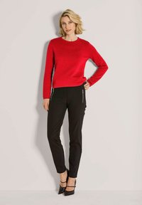 Frau in strukturiertem roten Pullover und schwarzen Hosen mit Reißverschlussdetails, die an einer neutralen Wand steht und schwarze spitze High Heels trägt.