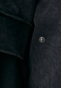 Schwarzer Stoff mit weicher Textur, ausgestattet mit einer sichtbaren silbernen Druckknopf und einem Kragen. Die Nähte sind dezent und passen zur Stofffarbe.
