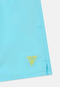 Pantaloni da bagno turchesi con una texture liscia, caratterizzati da una toppa triangolare gialla con il logo "GUESS" e un design di palma.