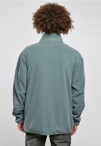 Fleece-pullover i dæmpet teal med høj krave, der har grå detaljer på ærmerne og en afslappet, oversized pasform.