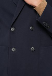 Σκούρο μπλε σακάκι διπλής σειράς με υφή, διαθέτει τέσσερα μαύρα κουμπιά και τσέπη στο στήθος. Μανίκια με μπάσκα και ανοιχτό καφέ περιθώριο.