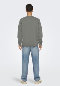 Hombre de pie de espaldas vistiendo una sudadera gris, vaqueros holgados de color azul claro y zapatos beige sobre un fondo blanco liso.